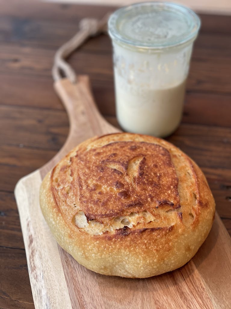 Freshly baked sourdough bread on a wooden board with a jar of sourdough starter.
