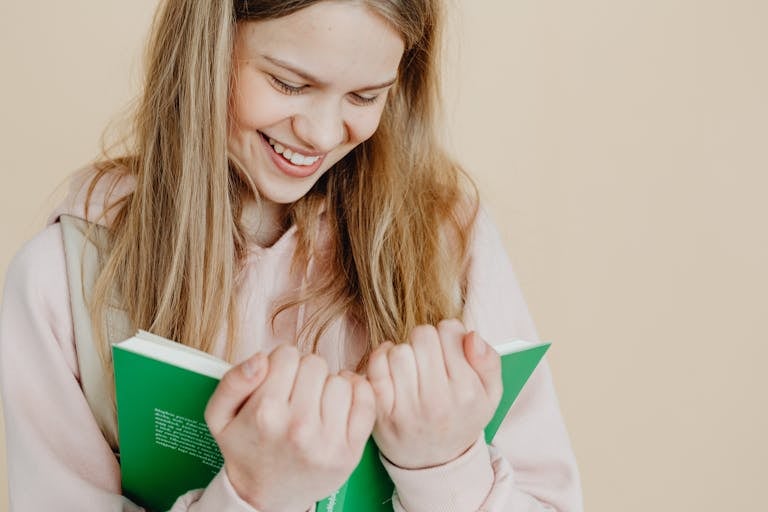 Happy teenager holding a green book against a beige background indoors.