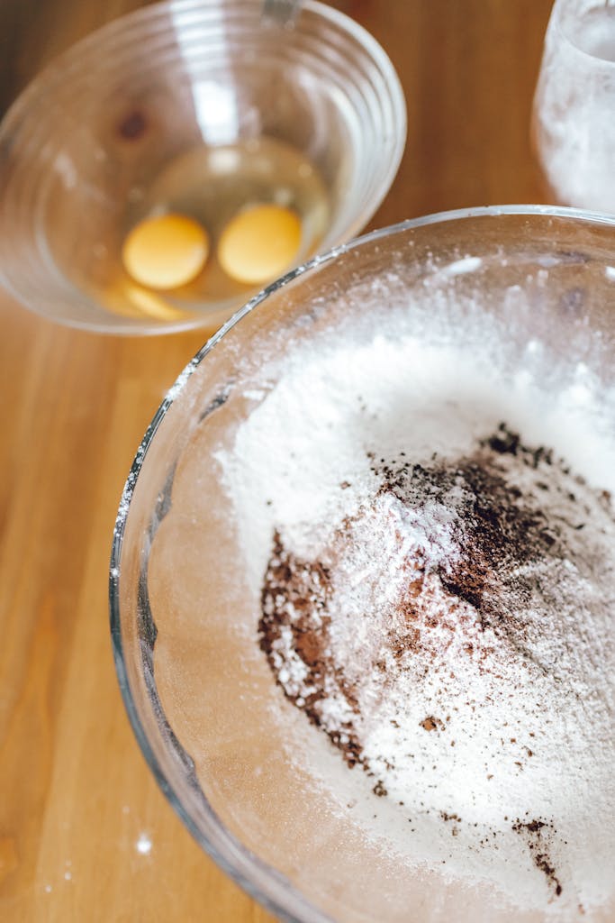 A close-up photo of flour, cocoa, and eggs in glass bowls on a wooden table.