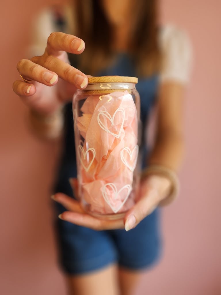A person holding a glass jar with pink paper and heart decorations, perfect for gifting.