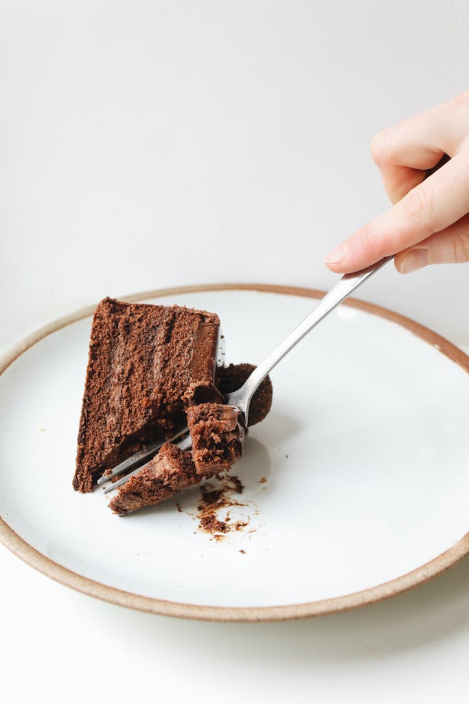 Close-up of a hand using a fork to slice a piece of chocolate cake on a plate.