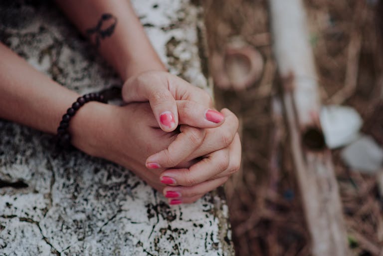 Close-up of hands clasped together adorned with pink nail polish, bracelet, and tattoo outdoors.