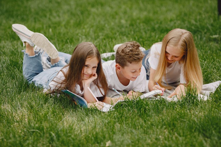Three children lying on grass reading and smiling outdoors.