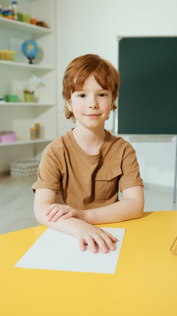 A happy child sitting at a table in a classroom, smiling warmly at the camera.