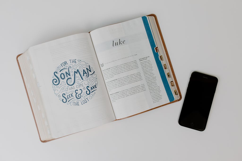 Open Bible displaying the Book of Luke next to a smartphone on a white surface.