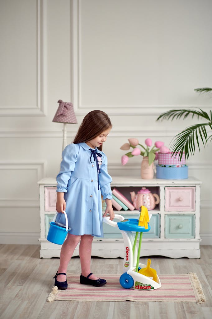 A young girl in a blue dress happily plays with a toy cleaning set inside a stylish room.