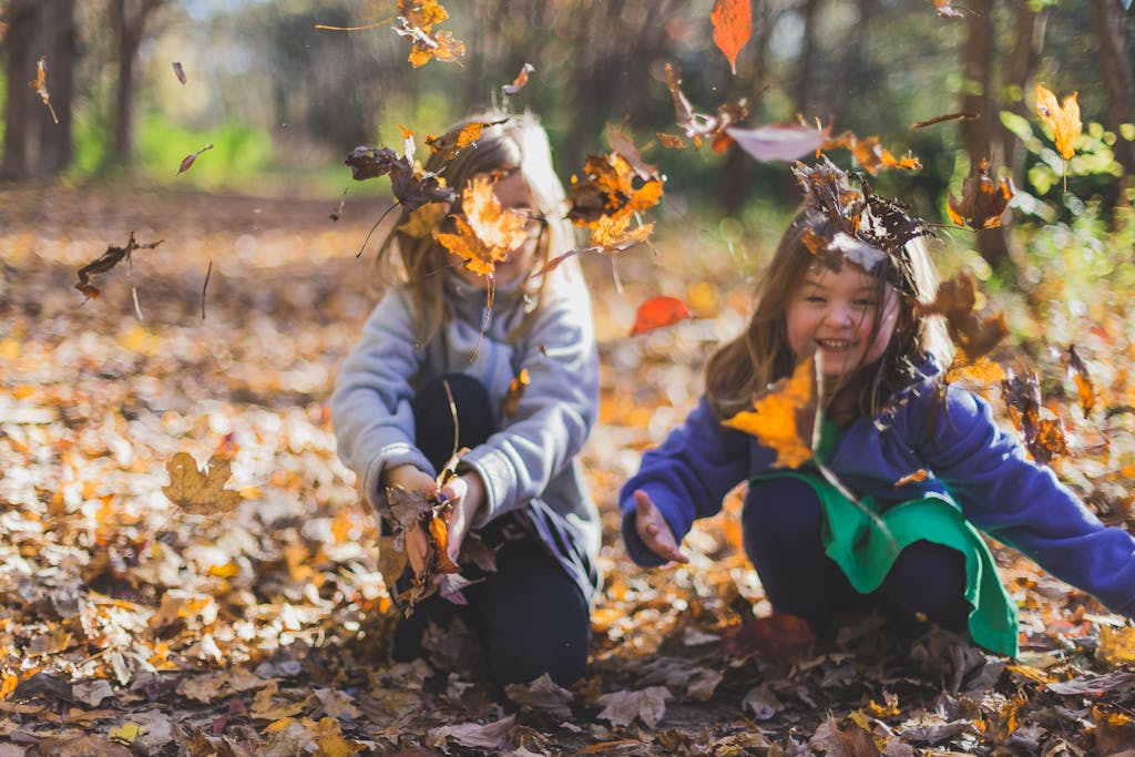 Two smiling girls playing joyfully amidst colorful fall leaves in a sunlit forest