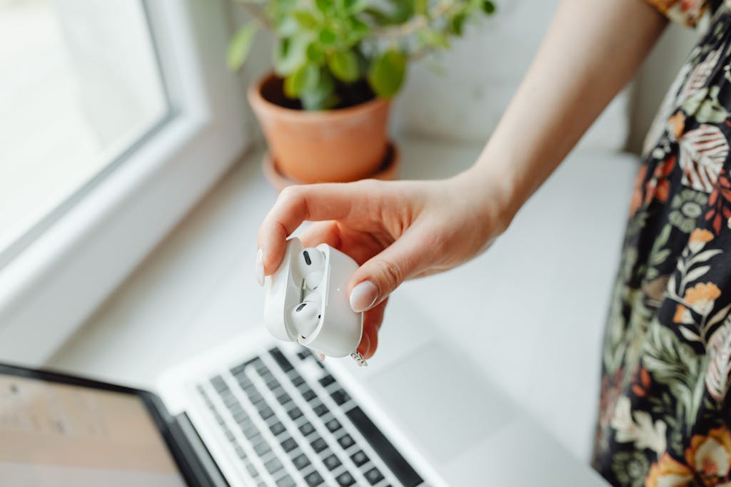 A person holds wireless earbuds over a laptop in a cozy home setting, with natural light from a window.