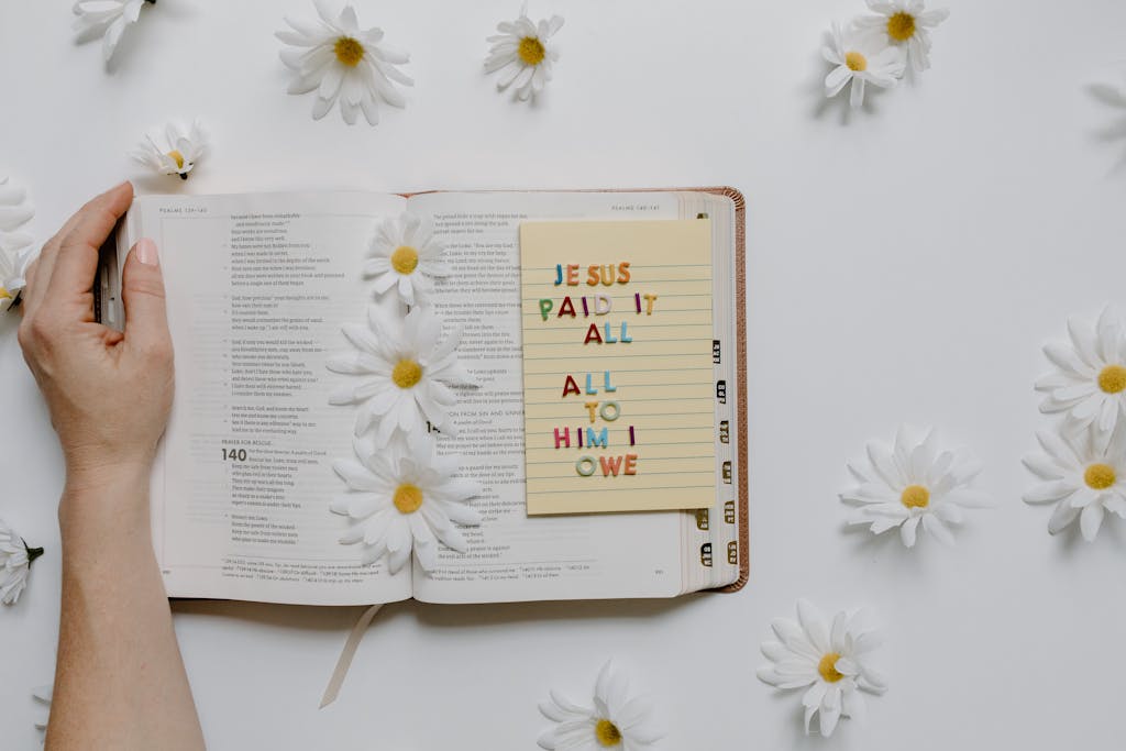 Open Bible with daisies and a note saying 'Jesus paid it all' creating a peaceful setting.